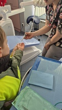 W gabinecie stomatologicznym dentystka pokazuje dziecku narzędzie dentystyczne, trzymając je nad jego dłonią. Dziecko w z zaciekawieniem wyciąga ręce. W tle widoczny jest fotel stomatologiczny oraz wyposażenie gabinetu.