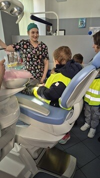 W gabinecie stomatologicznym dentystka w kolorowym stroju medycznym rozmawia z grupą dzieci. Jedno z dzieci siedzi na fotelu stomatologicznym, a pozostałe stoją obok i uważnie obserwują. Wokół widoczny jest sprzęt dentystyczny, w tym lampa zabiegowa i stanowisko pracy stomatologa.