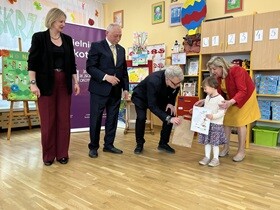 Dziewczynka w białym swetrze i sukience trzyma w dłoni dyplom. Przed nią nachyla się pan i wręcza brązową torbę. Obok stoi pan i pani.