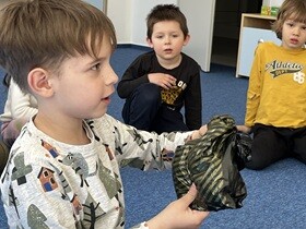Chłopiec w jasnej bluzce trzyma w dłoniach czarną torebkę. Za nim siedzą dwaj chłopcy.
