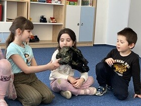 Troje dzieci siedzi na dywanie. Dziewczynka z lewej strony podaje czarną torebkę dziewczynce po prawej stronie.