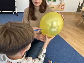 Kobieta trzyma w dłoniach nadmuchany żółty balon. Chłopiec rysuje na balonie niebieskim flamastrem kreskę.
