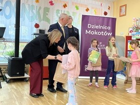Dziewczynka w różowej bluzce trzyma dyplom. Drugą ręką odbiera od pani torbę.