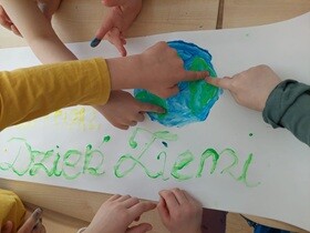 Dzieci malują farbami planetę Ziemię na dużym arkuszu papieru: kilka małych rączek, które wspólnie tworzą niebiesko-zielony glob, a obok widnieje napis „Dzień Ziemi” wykonany zieloną farbą.