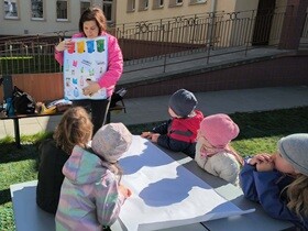 Nauczycielka prowadzi zajęcia na świeżym powietrzu, pokazując dzieciom planszę z kolorowymi pojemnikami do segregacji odpadów. Przedszkolaki siedzą przy stole i z uwagą słuchają, ucząc się zasad dbania o środowisko. W tle widoczny jest budynek przedszkola i plac zabaw.