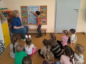 Nauczycielka prowadzi zajęcia edukacyjne w przedszkolu na temat oszczędzania wody. Dzieci siedzą na dywanie i z uwagą słuchają, a jedno z nich wskazuje elementy na planszy edukacyjnej przedstawiającej sposoby dbania o środowisko.