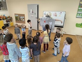 Dzieci stoją obserwując prowadzącą, która przebrana jest za Królową Matematykę.