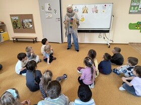Dzieci siedzą na dywanie. Przed nimi stoi nauczycielka przebrana za Królową Matematykę. W ręku trzyma liczydło.
