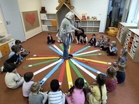 Prowadząca przebrana za Królową Matematykę stoi wewnątrz wiatraka matematycznego, który składa się z kolorowych szarf. Dzieci siedzą przed szarfami.