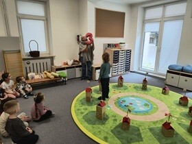 Dzieci siedzą w sali. Przed nimi leży okrągły zielony dywan, na którym namalowane są figury i ustawione drewniane domki. Przed dziećmi stoi prowadząca, która trzyma uniesioną, pluszową cyfrę 3.