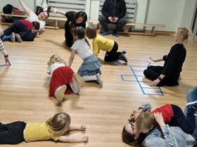 W sali z drewnianą podłogą trwa dynamiczna zabawa ruchowa z udziałem dzieci i dorosłych. Kilkoro dzieci porusza się po podłodze – czołgają się, leżą lub klęczą, angażując się w ćwiczenia. Na środku kobieta prowadząca zajęcia pochyla się nad dziećmi, aktywnie je wspierając i kierując zabawą.