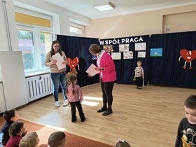 Dziewczynka podaje rękę pani dyrektor i odbiera dyplom i nagrody. Obok stoi nauczycielka trzymająca duży wiklinowy kosz.