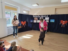 Chłopiec podaje rękę pani dyrektor i odbiera dyplomy i nagrody. Obok stoi nauczycielka trzymająca duży wiklinowy kosz. W tle widać troje innych nagrodzonych uczestników.