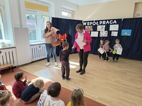 Chłopiec podaje rękę pani dyrektor i odbiera dyplomy i nagrody. Obok stoi nauczycielka trzymająca duży wiklinowy kosz. W tle widać troje innych nagrodzonych uczestników.