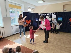 Dziewczynka podaje rękę pani dyrektor i odbiera dyplom i nagrody. Obok stoi nauczycielka trzymająca duży wiklinowy kosz. W tle widać dwoje innych nagrodzonych uczestników.