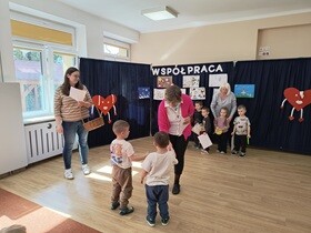 Dwoje chłopców odbiera dyplomy i nagrody od Pani Dyrektor. Jeden z chłopców podaje Pani Dyrektor dłoń. Obok stoi nauczycielka trzymająca duży wiklinowy kosz. W tle stoi 4 dzieci, które w rękach trzymają nagrody i dyplomy.