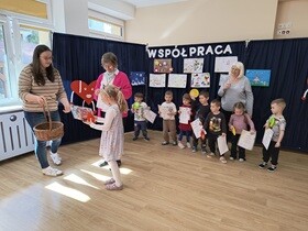 Siedmioro dzieci stoi obok siebie i w rękach trzyma dyplomy i nagrody. W tle widać granatowe płótno, na którym umieszone jest napis współpraca oraz przywieszone są prace konkursowe wykonane przez dzieci i rodziców. Dziewczynka odbiera nagrodę od nauczycielki i pani dyrektor.