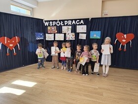Ośmioro dzieci stoi obok siebie i w rękach trzyma dyplomy i nagrody. W tle widać granatowe płótno, na którym umieszone jest napis współpraca oraz przywieszone są prace konkursowe wykonane przez dzieci i rodziców.