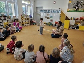 Dzieci siedzą w półkolu na dywanie. Przed nimi stoi tablica z napisem Wielkanoc oraz zdjęciami przedstawiającymi wybrane tradycje wielkanocne. Obok tablicy siedzi pani, która prowadzi zajęcia i rozmawia z dziewczynką stojącą przed tablicą.