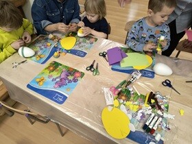 Przy stoliku siedzi 3 chłopców i kobieta. Wspólnie oblepiają jajka ze styropianu. Na stole znajdują się materiały kreatywne.