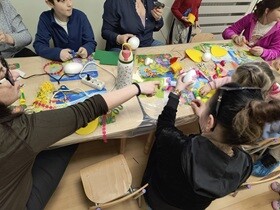 Przy stoliku siedzą dorośli wraca z dziećmi i wspólnie tworzą ozdoby wielkanocne.