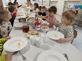 Dzieci siedzą przy stole, na którym znajdują się białe głębokie talerze, wazy, białe kubeczki na napoje, chleb, babka i dekoracje świąteczne. Jedno z dzieci trzyma w ręce chochelkę i nalewa sobie żurek z wazy na talerz.