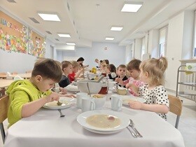 Dzieci siedzą przy stole, na którym znajdują się białe głębokie talerze, wazy, białe kubeczki na napoje, chleb, babka i dekoracje świąteczne. Jedzą świąteczny żurek.