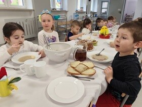 Dzieci siedzą przy stole, na którym znajdują się białe głębokie talerze, wazy, białe kubeczki na napoje, chleb, babka i dekoracje świąteczne.
