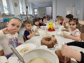 Dzieci siedzą przy stole, na którym znajdują się białe głębokie talerze, wazy, białe kubeczki na napoje, chleb, babka i dekoracje świąteczne.