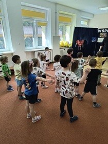 Dzieci stoją w rozsypce na dywanie i wyciągają w bok prawą rękę.
