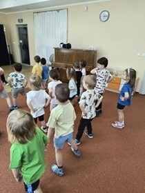Dzieci stoją twarzą do prowadzącego, trzymają się za boki i tańczą.