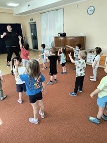 Dzieci stoją twarzą do prowadzącego i podnoszą ugięta lewą rękę.