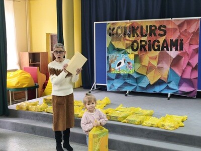 Dziewczynka trzyma w rękach torbę z nagrodami. Za nią stoi kobieta z dyplomem w ręku. Z tyłu jest kolorowy parawan z napisem konkurs origami i pracą wykonaną z origami. 