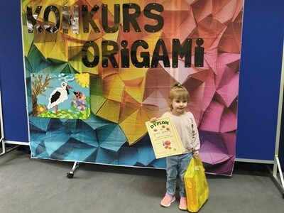 Dziewczynka trzyma w ręku dyplom a w drugiej rączce torbę z nagrodami. Za nią stoi kolorowy parawan z napisem konkurs origami i pracą wykonaną z origami.