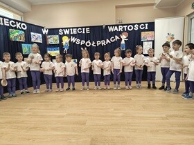 Dzieci stoją w półkolu i grają na instrumentach bębenkach, kołatkach, grzechotkach, klawesach. W tle widać granatowy parawan, na którym przyczepiony jest napis "Dziecko w świecie wartości", prace dzieci oraz postać chłopca i dziewczynki trzymających napis współpraca.