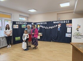 Dziewczynka odbiera nagrodę i stoi do zdjęcia z Panią Naczelnik oraz Panią Dyrektor. W tle widać granatowy parawan, na którym przyczepiony jest napis "Dziecko w świecie wartości", prace dzieci oraz postać chłopca i dziewczynki trzymających napis współpraca.