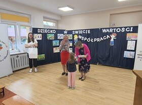Dziewczynka odbiera dyplom od Pani Dyrektor. Pani Naczelnik i nauczycielka biją brawo. W tle widać granatowy parawan, na którym przyczepiony jest napis "Dziecko w świecie wartości", prace dzieci oraz postać chłopca i dziewczynki trzymających napis współpraca. Obok stoi baner z logiem przedszkola.