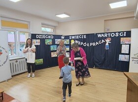 Chłopiec podchodzi do Pani Dyrektor i odbiera dyplom. W tle widać granatowy parawan, na którym przyczepiony jest napis "Dziecko w świecie wartości", prace dzieci oraz postać chłopca i dziewczynki trzymających napis współpraca.
