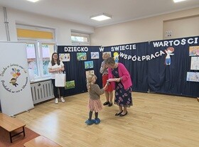 Dziewczynka stoi obok Pani Dyrektor i odbiera dyplom. W tle widać granatowy parawan, na którym przyczepiony jest napis "Dziecko w świecie wartości", prace dzieci oraz postać chłopca i dziewczynki trzymających napis współpraca.