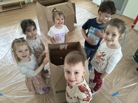 6 dzieci stoi naokoło domku z kartonu i uśmiecha się do zdjęcia.