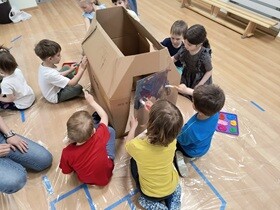 Dzieci siedzą na folii naokoło domku z kartonu i malują go farbami.