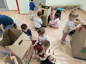 Dzieci siedzą lub stoją na folii naokoło domków z kartonu i malują je farbami.