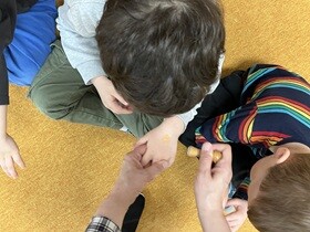 Dwóch chłopców siedzi na dywanie. Chłopiec w szarej bluzce ma wyciągniętą dłoń, na jej wierzchniej części widać pieczątkę.
