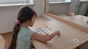 Dziewczynka przed lustrem, układa obrazki, rozłożone na blacie stolika.
