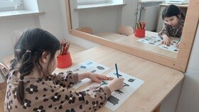 Dziewczynka siedzi przy stoliku przed lustrem, ma przed sobą dwie karty pracy z obrazkami i kredki w pojemniku. Kredką zaznacza na jednej karcie wybrane obrazki.