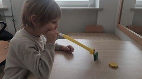 Chłopiec siedzi przy stoliku, jedną ręką trzyma rurkę przy buzi, w którą dmucha próbując przesunąć dwa przedmioty leżące na stoliku.