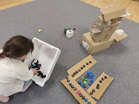 Dziewczynka kuca na dywanie mieszając dłonią w szufladzie z plastikowymi nakrętkami. Przed nią stoi budowla z kartonu oraz leży karton udekorowany korkami.
