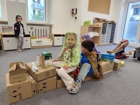 Dwie dziewczynki i chłopiec siedzą na pociągu zrobionym z ułożonych kartonów. Ubrania są w kolorowe chusty i apaszki.
