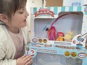 Dziewczynka stoi obok sceny/ teatrzyku zrobionego z dwóch kartonów. Bawi się zrobionymi z papieru kukiełkami. 