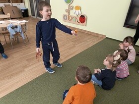 Dzieci z młodszej grupy siedzą na dywanie. Przed nimi stoi uśmiechnięty chłopiec, który pokazuje on potworka zrobionego z żółtego, małego opakowania po niespodziance.